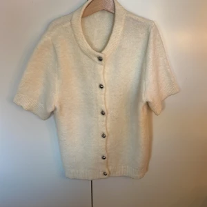 Vit kofta med kort ärm från Gina Tricot - Säljer en söt vit kofta från Gina Tricot i storlek XS. Koftan har korta ärmar, mjukt stickat material och knäpps med silvriga knappar framtill. Perfekt att styla till jeans eller kjol. Koftan har inga defekter och är använd ett fåtal gånger.
