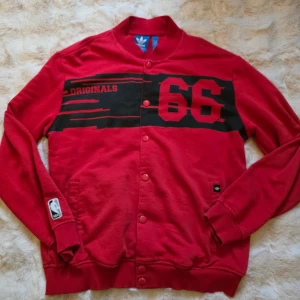 Röd Adidas Originals jacka Chicago Bulls  - Säljer en röd Adidas Originals jacka med tryck '66' och 'Originals' på bröstet. Jackan har knappar framtill, ribbade muddar och NBA-logga på ärmen. Klassisk streetstil med svart detalj över bröstet och Adidas-märke vid fickan.