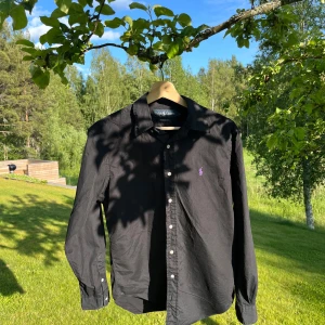 Svart skjorta från Ralph Lauren S - Klassisk svart skjorta från Ralph Lauren i bomull med lila broderad logga på bröstet. Skjortan har lång ärm, knappar framtill och klassisk krage. Passformen är classic fit och materialet är mjukt och skönt. Perfekt nu för sommaren! Det går att göra grymma outfits med den!
