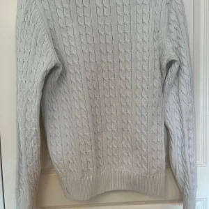 Ljusgrå kabelstickad tröja Holly & Whyte - Kabelstickad tröja i ljusgrått från Holly & Whyte by Lindex. Klassisk rund halsringning, ribbade muddar och lång ärm. Snyggt mönster med breda flätor och liten metallbricka vid nederkanten. Perfekt för lager-på-lager under kyliga dagar.