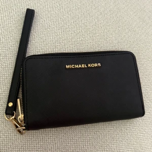 Svart plånbok från Michael Kors - Stilren svart plånbok från Michael Kors i skinn med guldfärgade detaljer och dragkedja runtom. Plånboken har flera kortfack, fönster för ID och ett myntfack med dragkedja. Praktisk handledsrem gör den enkel att bära med sig.