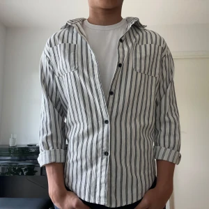 Randig vit skjorta med svarta knappar - Snygg vit skjorta med mörkblåa ränder och svarta knappar. Modellen är 184. 