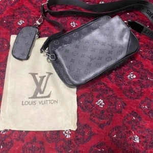Svart axelväska monogram - Säljer en svart axelväska med klassiskt monogrammönster. Väskan har bred justerbar axelrem, flera fack och en mindre avtagbar plånbok. Tillverkad i skinn med silverfärgade detaljer. Perfekt för dig som vill ha en stilren och lyxig accessoar.
