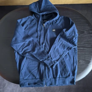 Blå hoodie från Under Armour - Snygg marinblå hoodie från Under Armour med dragkedja framtill och huva med snörning. Tröjan har långa ärmar, två fickor och en liten logga på bröstet. Perfekt för chill dagar eller till träningen.