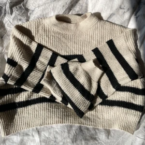 Beige och randig stickad tröja - Två stickade tröjor i beige toner, en med svarta ränder och en helt beige. Båda har ribbade muddar, hög hals och lång ärm. . Snyggt avslappnad passform och mjukt material, samt croppad 