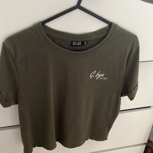 Olivgrön croppad t-shirt - Olivgrön croppad t-shirt med korta ärmar och rund hals. Tryck med texten 'G.bye xoxo' på bröstet. Skön och enkel modell i mjuk bomull, perfekt till jeans eller shorts. Ärmarna är lätt upprullade för en avslappnad vibe.