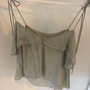 Zara Grön volangig blus  - Supersöt ljusgrön blus med tunna axelband som knyts och volang över bysten. Blusen är luftig och har pärlbroderi längs volangen för extra detalj. Perfekt till sommaren och ger en romantisk vibe.