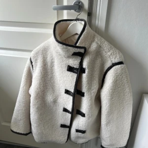 Vit teddyjacka med svarta detaljer - Säljer en riktigt snygg vit teddyjacka med svarta kantband och coola knäppningar framtill. Jackan har hög krage och en boxig, loose passform som ger en trendig vibe. Perfekt för dig som vill sticka ut med en unik och mysig jacka i vinter.