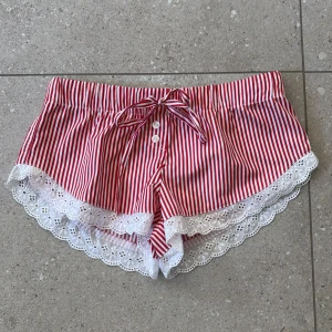 Randiga shorts med spets från Fanny Castelius - Söta shorts med röda och vita ränder från Fanny Castelius. De har knytband i midjan, knappar framtill och är dekorerade med vit broderad spets längs bensluten. Perfekta för sommarens varma dagar och ger en lekfull vibe.👩🏼‍❤️‍💋‍👨🏼🫶🏻💗