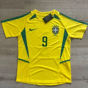 Brasilien Ronaldo 9 Nike fotbollströja - Gul Brasilien fotbollströja från Nike med gröna detaljer, nummer 9 och Ronaldo på ryggen. Klassisk CBF-logga broderad på bröstet och grönt Nike-märke. Tillverkad i lätt och ventilerande polyester, perfekt för match eller träning.
