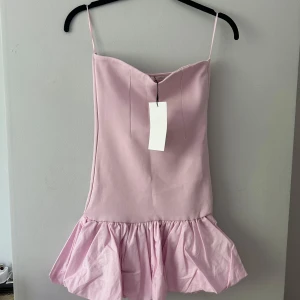 Ljusrosa volangklänning från Zara - Ny med prislapp!Supersöt ljusrosa kort klänning från Zara med tunna axelband och volang nertill. Klänningen har en enkel, rak passform upptill och en fluffig volangkjol som ger en lekfull vibe. Perfekt för dig som vill sticka ut med en feminin och trendig look.