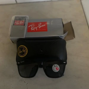 Svarta Ray-Ban Wayfarer solglasögon - Snygga svarta Ray-Ban Wayfarer solglasögon med polariserade glas. Klassisk fyrkantig form och diskret logga på skalmen. Kommer med originalfodral och box. Perfekta för att ge din stil en cool edge i solen.