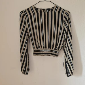 Randig croppad blus från Gina Tricot - Snygg croppad blus från Gina Tricot i svart och beige med vertikala ränder. Modellen har långa ärmar, rund halsringning och smockad resår i midjan. Stängs med knapp i nacken. Perfekt för dig som gillar trendiga detaljer och stilrena mönster.
