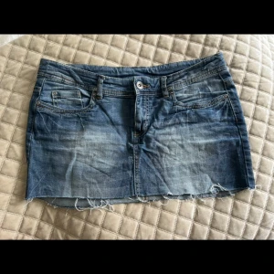 Blå jeanskjol med rå kant - Snygg blå jeanskjol med slitna detaljer och rå, fransig kant nertill. Klassisk femficksmodell med bälteshällor och dragkedja fram. Perfekt för en avslappnad och trendig look.