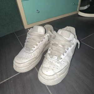 Vita Nike Air Force 1 sneakers - Klassiska vita Nike Air Force 1 sneakers med låg profil och rund tå. Skorna har snörning och är tillverkade i skinn med perforerade detaljer på tån för extra ventilation. Perfekta för en streetwear-look och passar till det mesta.