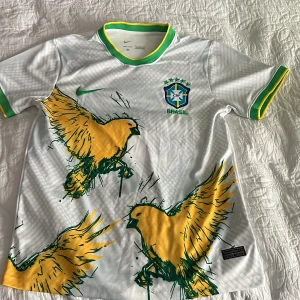 Vit Brasilien fotbollströja Nike S - Snygg vit Brasilien fotbollströja från Nike i Dri-FIT-material. Tröjan har korta ärmar, gröna och gula detaljer på krage och ärmslut samt stora gula fåglar tryckta framtill. Brasilien-emblem på bröstet och Nike-logga. Perfekt för dig som älskar fotboll och vill sticka ut.T-shirten är ej andvänd