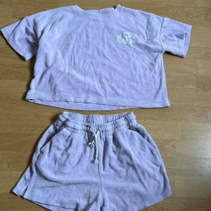 Ljuslila frottéshorts med tillhörande tröja  från Lindex - Mysiga ljuslila shorts och tröja från Lindex i mjuk frotté. De har elastisk midja med snörning och praktiska fickor. Perfekta för chill dagar eller efter badet. Matchar toppen med broderad text 'Ray of Sunlight'. Båda är i storlek 158-164 men passa även lite större 