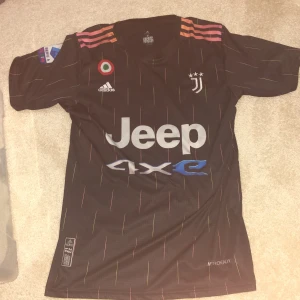Juventus Ronaldo 7 svart matchtröja - Juventus fotbollströja med Ronaldo 7 på ryggen. Svart bas med tunna färgade linjer, rosa och orange detaljer på axlarna, officiella loggor och sponsortryck. Tillverkad i lätt funktionsmaterial (AEROREADY) för bästa komfort på planen.