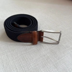 Mörkblått flätat bälte  - Stilrent mörkblått flätat bälte från Saddler med detaljer i brunt skinn och silverspänne. Bältet har en klassisk design med snygg kontrast mellan det svarta bandet och de bruna skinnpartierna. Perfekt accessoar för att lyfta din outfit. Hör av dig med frågor