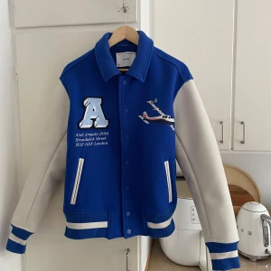 Blå och Vit Axel Arigato Jacka - Snygg blå och vit bomberjacka från Axel Arigato, flygplansmotiv och text på bröstet. Jackan har vita ärmar, ribbade muddar och knappar framtill. Stor logga på ryggen. Perfekt statement-plagg för dig som vill sticka ut. Nypris på jackan är 4000kr men den går inte att köpa ny längre. Därav ännu mer exklusiv. Passar Strl M då den är lite stor i strl.