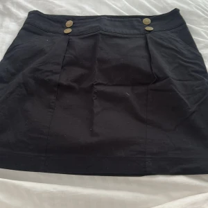 Svart kortkjol med knappar - Svart kortkjol med två dekorativa guldknappar framtill. Kjolen har en enkel, rak passform och är sydd i ett slätt tyg. 