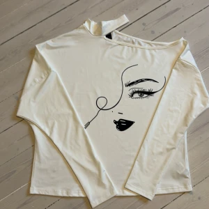 Vit långärmad topp med ansiktsmotiv - Vit långärmad topp i mjukt material med ett stilrent svart tryck av ett ansikte framtill. Tröjan har en något lös passform och bred halsringning, vilket ger en chill och modern vibe. Perfekt för dig som gillar minimalistisk design med en twist. 
