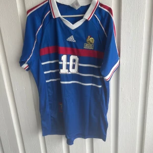Frankrike Zidane 10 Adidas fotbollströja - Klassisk blå fransk landslagströja från Adidas med nummer 10 och Zidane på ryggen. Tröjan har vita och röda detaljer, broderad tupp-logga och vita ränder över bröstet. Ribbad vit krage med röd och blå kant. Perfekt för fotbollsfans!