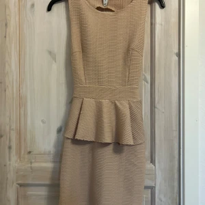 Beige peplumklänning med öppen rygg - Snygg beige kortklänning med peplumdetalj i midjan och cool öppen rygg med korsade band. Klänningen har en strukturerad, ribbad yta och är ärmlös med rund halsringning framtill. Perfekt för dig som vill sticka ut med en unik look.