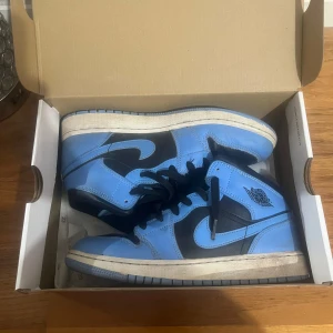 Nike Air Jordan 1 Mid blå/svarta - Säljer ett par Nike Air Jordan 1 Mid i blått och svart skinn med klassisk vinge-logga på sidan. Skorna har svarta snören, perforerad tå och vit sula. Perfekta för dig som vill sticka ut med en ikonisk sneaker i snygg färgkombo.