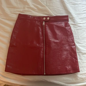 Röd kort kjol i skinnlook från Divided - Snygg röd kort kjol från Divided by H&M i glansig skinnimitation. Kjolen har en rak passform, silverfärgad dragkedja framtill och två tryckknappar i midjan. Perfekt statement piece för att lyfta din outfit.
