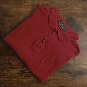 Fred Perry Pikétröja  - Fred Perry Green Label piké i vinröd färg, storlek 46 (M). Stilren och klassisk modell med ton-i-ton brodyr. Några små hål finns, men i övrigt bra skick. Perfekt för dig som gillar vintage och casual stil. Green Label – made in England! (3 små hål)