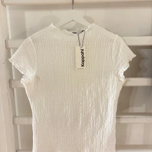 Vit topp från Kappahl - Superfin vit topp från Kappahl med smockad struktur och lätt volang vid ärmslut och halsringning. Kortärmad och i stretchigt material.