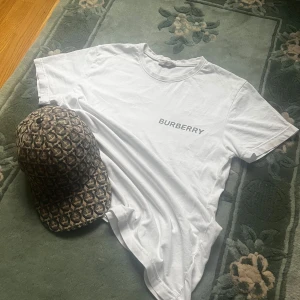 Vit Burberry t-shirt med stor logga - Vit t-shirt från Burberry i bomull med klassisk rund hals och korta ärmar. Framsidan har en stilren Burberry-text och baksidan pryds av en stor B-logga i blått och vitt mönster. Perfekt för dig som gillar exklusiva streetwear-vibbar.