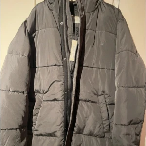 Grå pufferjacka från Rosemunde - Snygg grå pufferjacka från Rosemunde med quiltad design och dragkedja framtill. Jackan har huva, stora fickor och långa ärmar. Perfekt för kalla dagar när du vill hålla stilen och värmen. Klassisk look som funkar till allt.