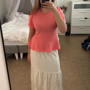 Korallrosa blus med volangkant - Supersöt korallrosa blus från Lindex med korta puffärmar och volangkant nedtill. Blusen har ett diskret hålmönster över hela tyget och en liten öppning med knapp i nacken. Perfekt till sommaren! Aldrig använt, prislappen är kvar.