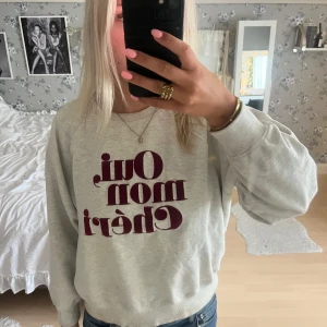Grå sweatshirt med vinröd text - Säljer en ljusgrå sweatshirt med trycket 'Oui mon chéri' i vinrött framtill. Tröjan har rund hals, långa ärmar och en relaxed passform. Perfekt för dig som gillar statement-prints och en chill vibe.