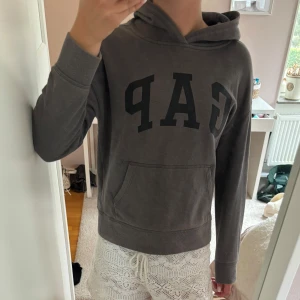 Mörkgrå GAP hoodie med huva - Säljer en mörkgrå hoodie från GAP med stor svart logga framtill. Tröjan har huva, känguruficka och långa ärmar. Tröjan är i barnstorlek men skulle säga att den passar en XS. (Obs dammtussen på bild 3 är från spegeln och ej tröjan) Skriv för frågor eller bilder! 