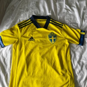 Sverige fotbollströja Adidas gul/blå - Gul Sverige fotbollströja från Adidas med blå detaljer och klassiska tre ränder på axlarna. Broderat landslagsmärke på bröstet och Adidas-logga. Tillverkad i lätt, ventilerande funktionsmaterial. Perfekt för match eller träning.