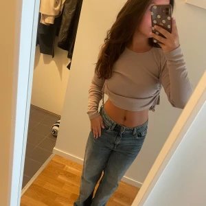 Beige croppad långärmad tröja Nelly - Säljer en beige croppad långärmad tröja med rund hals och snygga snörningsdetaljer i sidorna. Tröjan har en tight passform och är tillverkad i ett ribbat material som känns mjukt mot huden. Perfekt att matcha med jeans för en chill look.