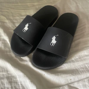 Svarta sandaler från Polo Ralph Lauren - Svarta slides från Polo Ralph Lauren med klassisk logga i vitt på ovandelen. Tillverkade i syntetmaterial med räfflad sula för extra grepp. Perfekta för chill dagar eller att slänga på sig till stranden.