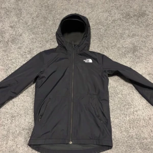 Svart vindjacka från The North Face - Svart vindjacka från The North Face med huva och hel dragkedja framtill. Jackan har två sidofickor med dragkedja och logga på både bröstet och ryggen. Perfekt för blåsiga dagar och enkel att matcha med allt.