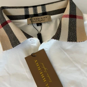 Vit Burberry t-shirt med rutig krage - Vit t-shirt från Burberry med klassisk rutig krage i beige, svart och rött. T-shirten har korta ärmar och knappar vid halsen. Snygg och stilren design med exklusiv känsla, perfekt för dig som gillar premium streetstyle.