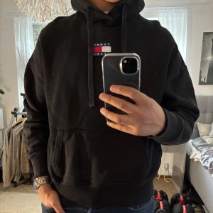 Svart hoodie Tommy Jeans med logga - Svart hoodie från Tommy Jeans med klassisk broderad logga på bröstet. Tröjan har huva med snörning, känguruficka och ribbade muddar. Perfekt för dig som gillar en clean och stilren look. Materialet är mjuk bomull som känns skönt mot huden. (S men väldigt oversized så passar för M och L med)
