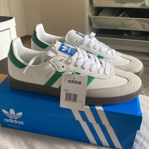 Adidas Samba OG vit/grön sneakers - Adidas Samba OG sneakers i vitt skinn med gröna detaljer och klassiska tre ränder på sidan. Grå mocka vid tån, brun gummisula och vit snörning. Ikonisk retrostil med blå Adidas-logga på plösen. Perfekta för dig som gillar streetwear och tidlös design.