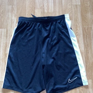 Nike Dri-Fit svarta sportshorts L/XL - Svarta sportshorts från Nike med vita sidor och broderad logga framtill. Tillverkade i Dri-Fit polyester som andas, med elastisk midja och snörning för bästa passform. Perfekta för träning eller sportiga dagar.