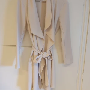 Beige trenchcoat från ONLY, storlek L - Snygg beige trenchcoat från ONLY med långa ärmar och bälte i midjan. Jackan har en omlott-design med breda slag och ett flowigt fall. Perfekt för dig som gillar stilrena och trendiga plagg med en feminin touch.