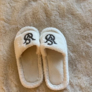 Vita fluffiga tofflor med glitterdetalj - Supermysiga vita tofflor i fluffigt syntetmaterial med svart glittrande logga på ovansidan. De har öppen tå och platt sula, perfekta att glida runt i hemma. Snygg och bekväm design som ger en lyxig känsla till vardagen.