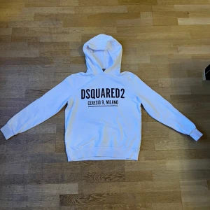 Vit hoodie från Dsquared2 med tryck - Säljer en snygg vit hoodie från Dsquared2 med svart tryck på bröstet där det står 'DSQUARED2 Ceresio 9, Milano'. Bild finns! Tröjan har huva, långa ärmar och klassisk passform. Perfekt för dig som gillar streetwear och vill sticka ut med ett stilrent plagg.