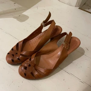 Bruna skinnpumps från Chie Mihara - Snygga bruna pumps i skinn från Chie Mihara med öppen tå och flätad detalj framtill. Skorna har låg klack, justerbar rem med spänne och klassisk retrovibe. Perfekta för dig som gillar stilrena och tidlösa skor med en twist.