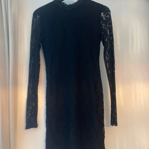 Svart spetsklänning med lång ärm - Supersnygg svart kortklänning i spets med långärmade ärmar och hög hals. Klänningen har ett blommigt spetsmönster och dragkedja bak. Bra skick.